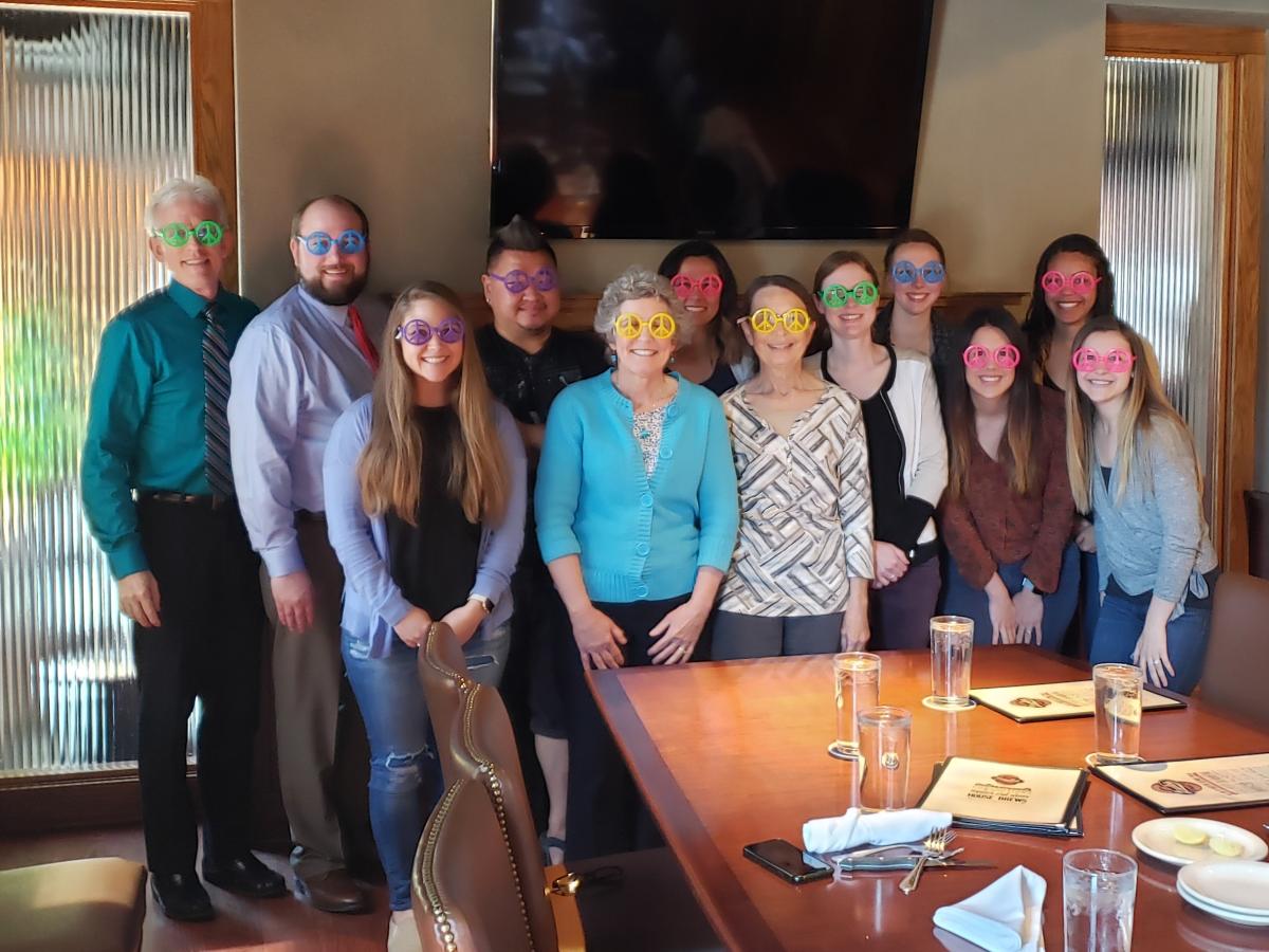 A group photo of Dave Hansen's lab. In the back row is Dave Hansen, Matt Hansen, Zach Huit, Akemi Mii, Jessie Tibbs, and Kelsey McCoy. In the front row is Brittany Sutton, Mary Kay Hansen, Mary Fran Flood, Katie Meidlinger, Emily Sonnen, and Kate Theimer. They are all wearing party glasses shaped like peace signs in various neon colors.