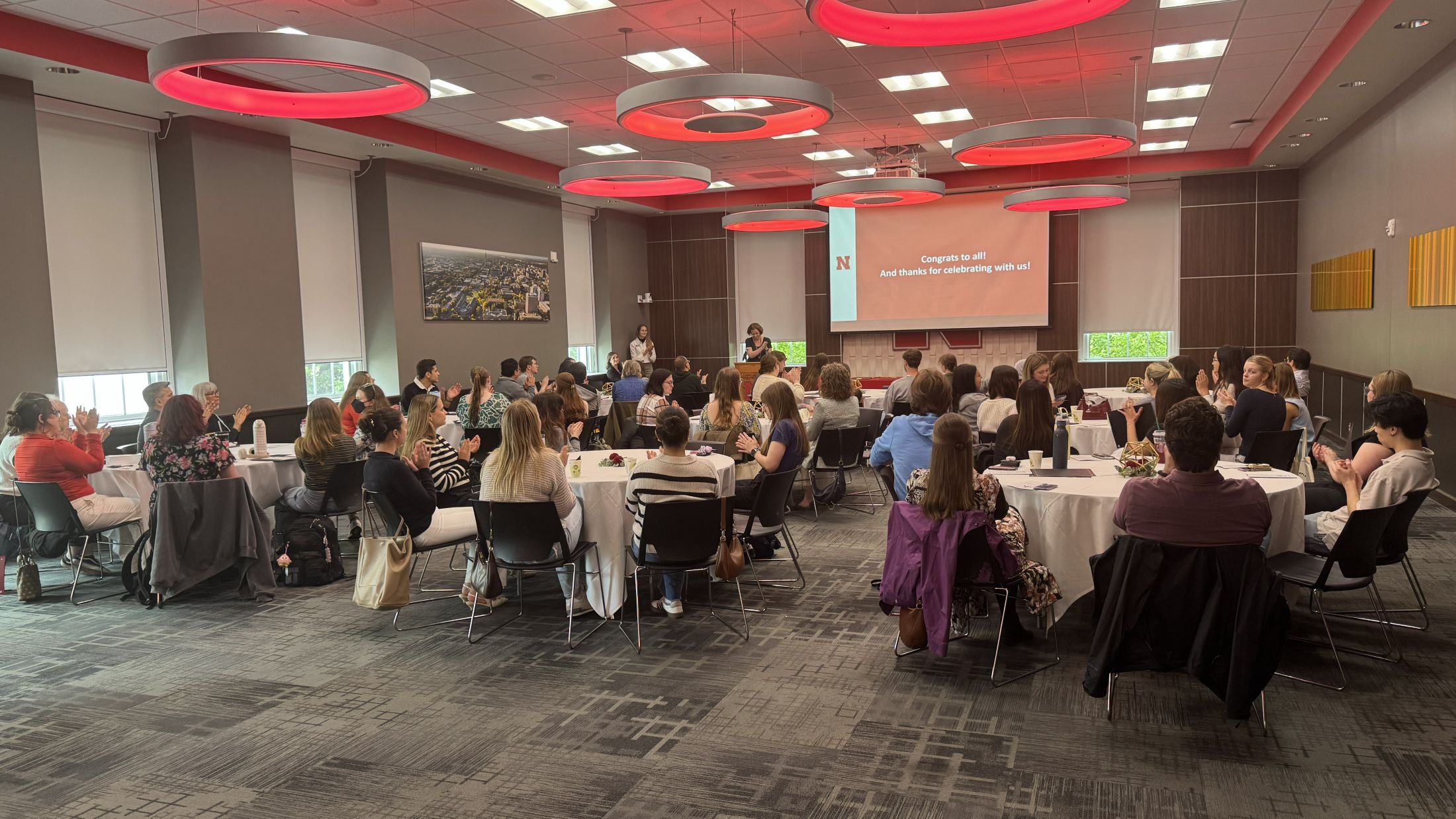 Room with people seated at round tables, facing presenters near a screen in a modern conference setting.