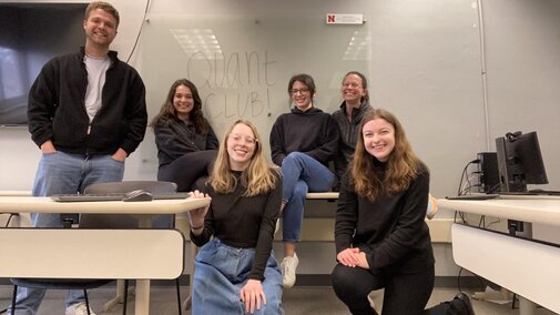 Quant Club students sitting and standing in a classroom with Quant CLUB! written on the dry-erase board behind them.